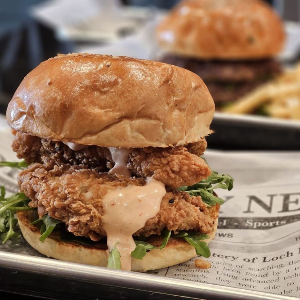 A Chick N Burger fried chicken sandwich with sauce and arugula on a bun, with another burger in the background.