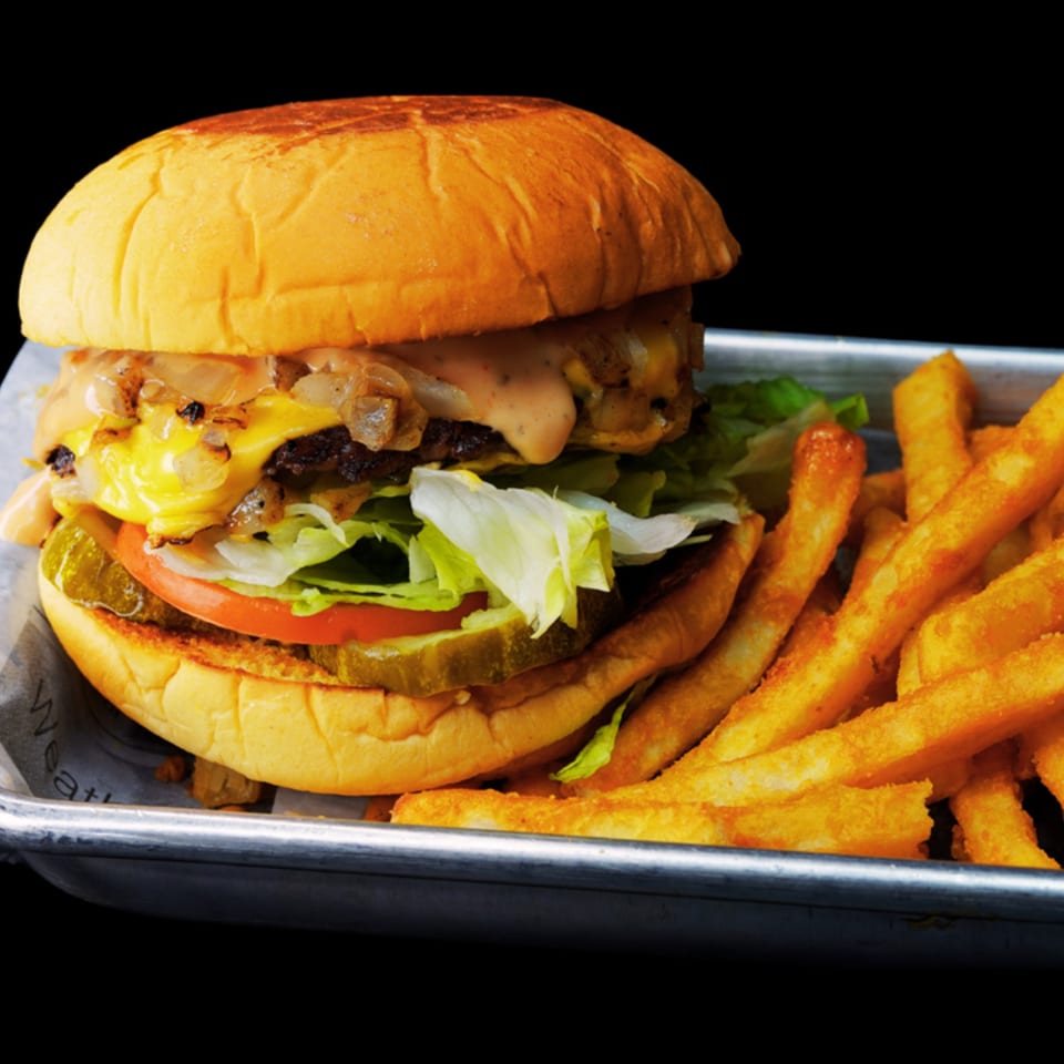 A Chick N Burger cheeseburger with lettuce, tomato, pickles, and fries on a metal tray.